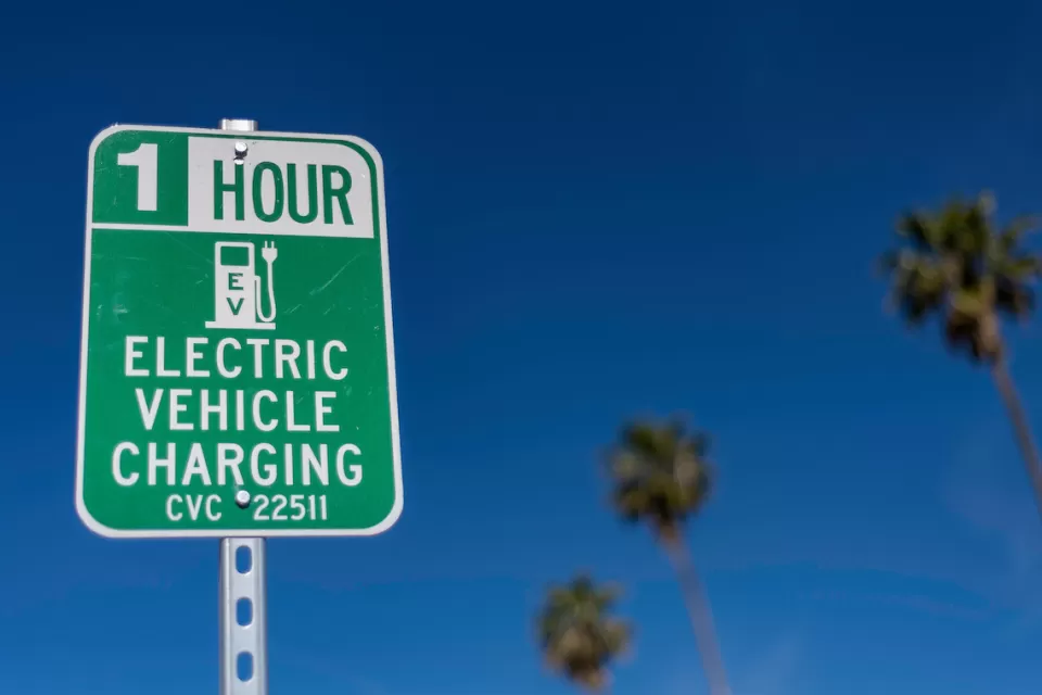 Close-up of green and white sign for 1 hour EV charging station outdoors with tall palm trees in background against blue sky.